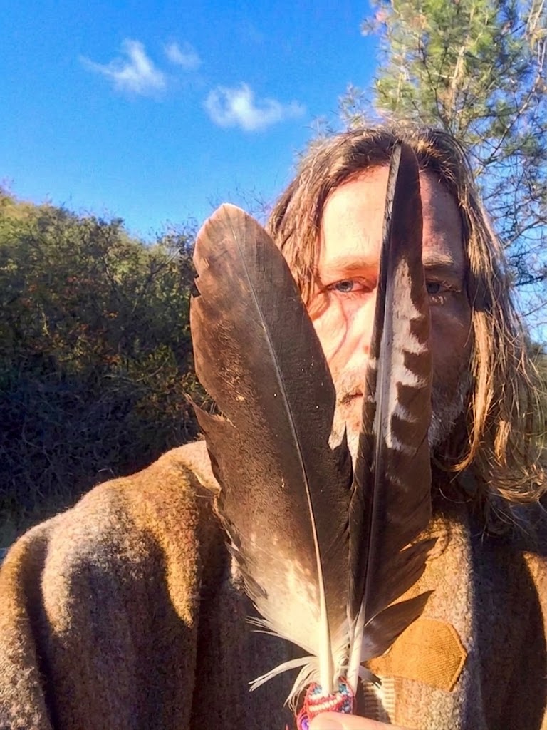 Lore Solaris holding two eagle feathers to his face, in a woollen cloak under a blue sky